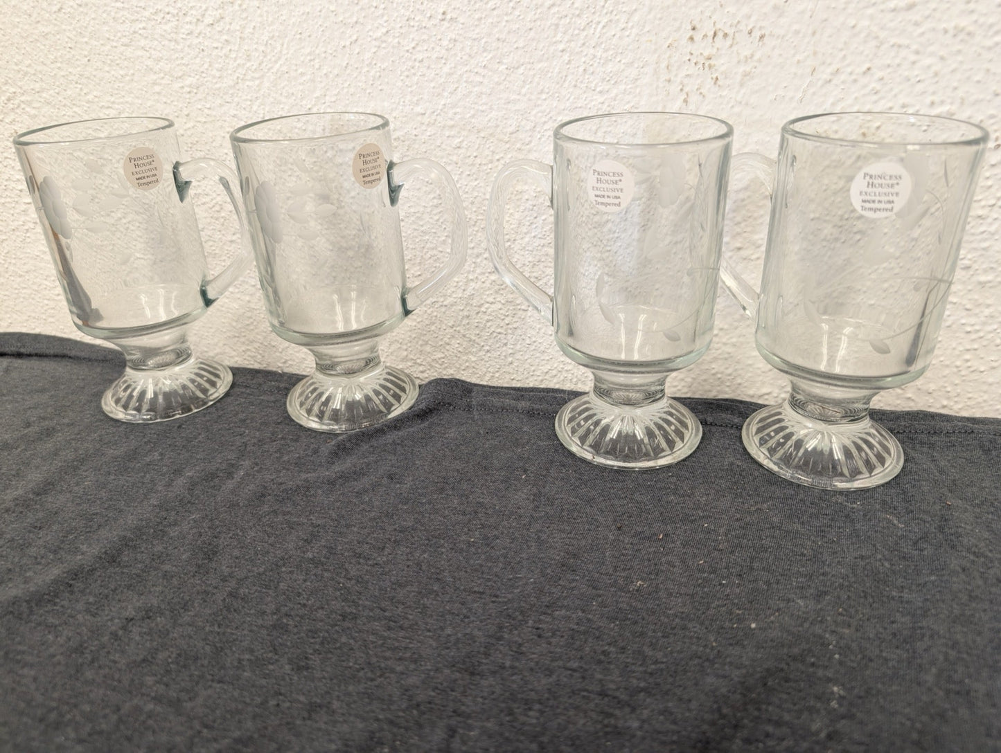 Four clear glass pedestal mugs lined up, featuring delicate floral etching and foil stickers.