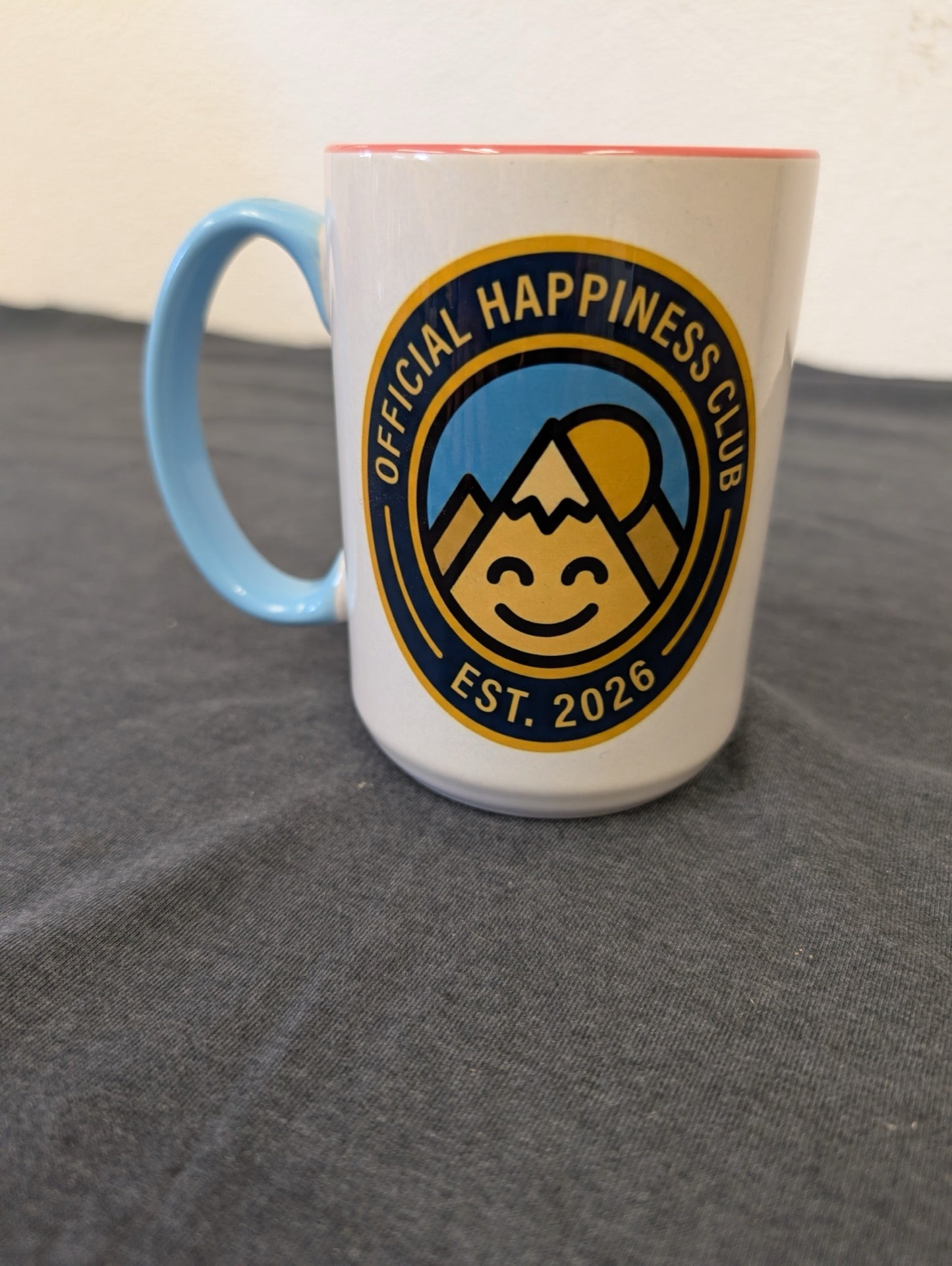 White mug with a blue handle and a yellow/blue logo reading Official Happiness Club Est. 2026.