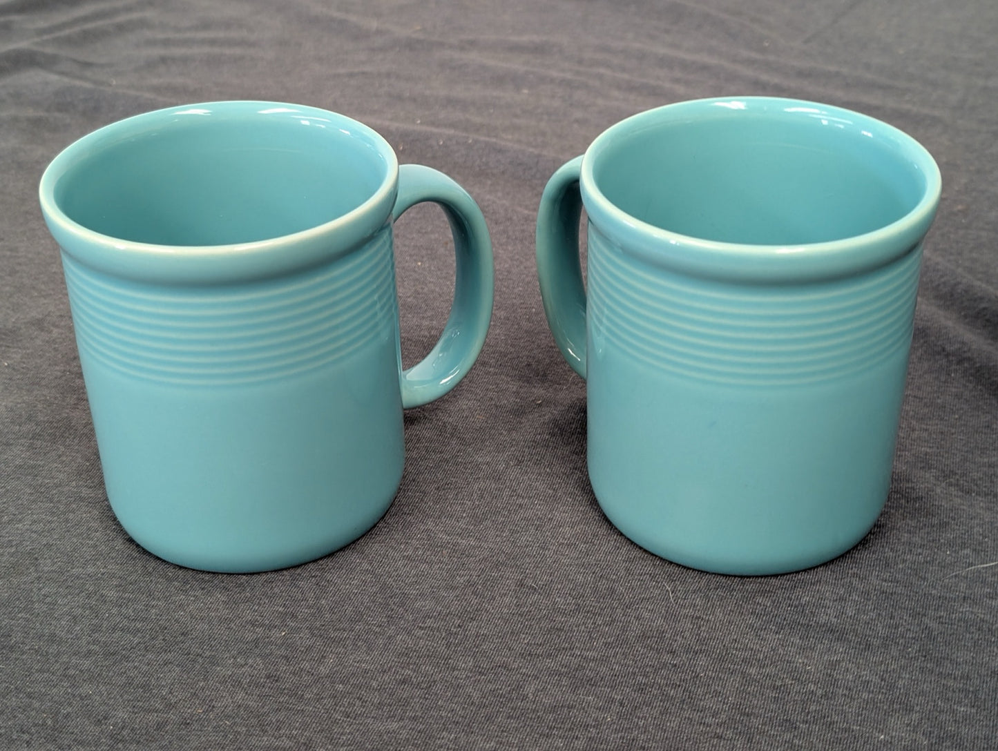 Two identical teal blue ceramic mugs with horizontal ribbed detailing on a gray background.