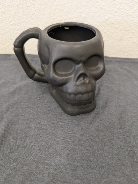 Matte black ceramic coffee mug shaped like a human skull with a bone handle.