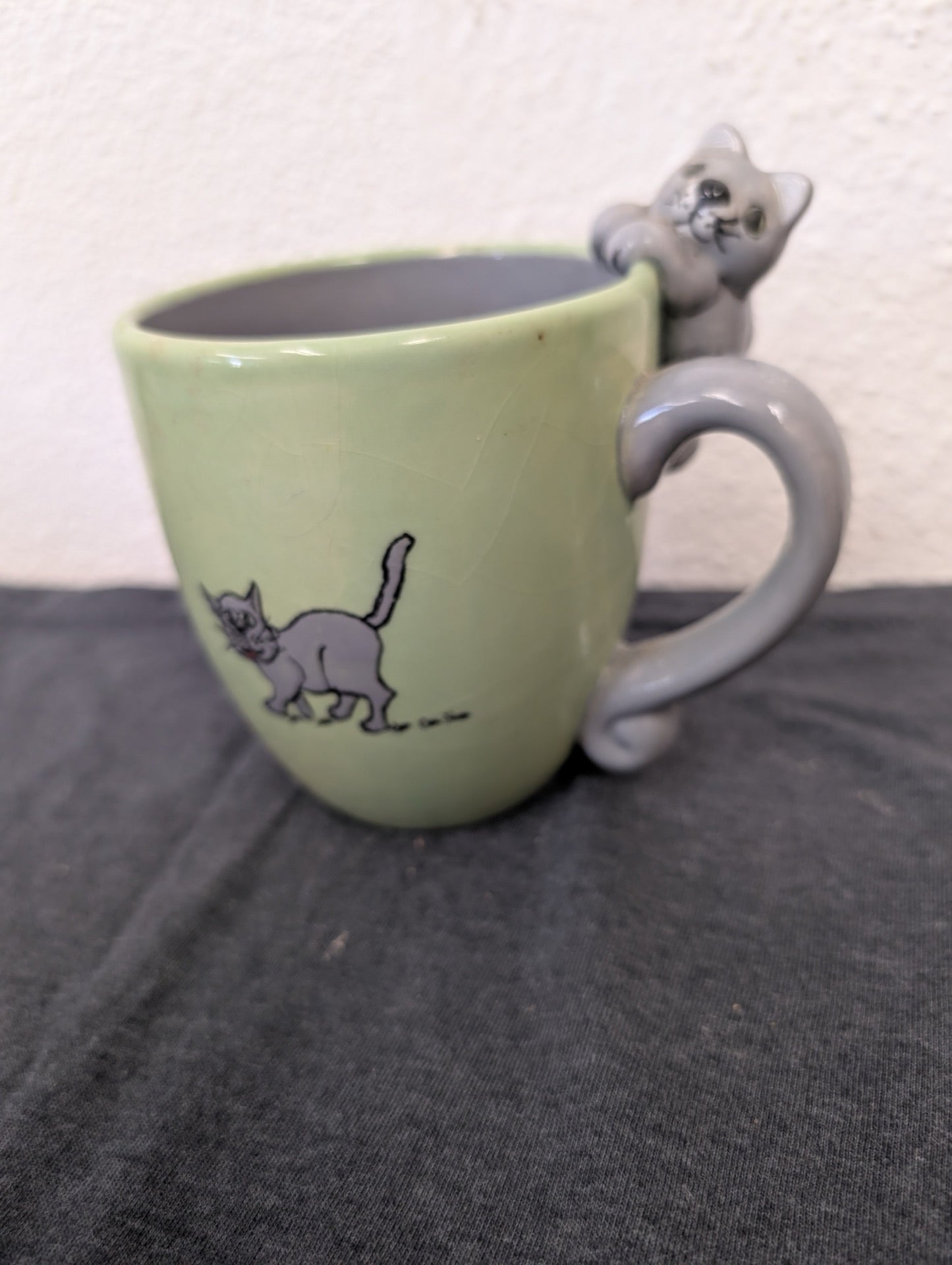 Light green ceramic mug featuring a drawing of a cat and a 3D cat figure forming the handle.