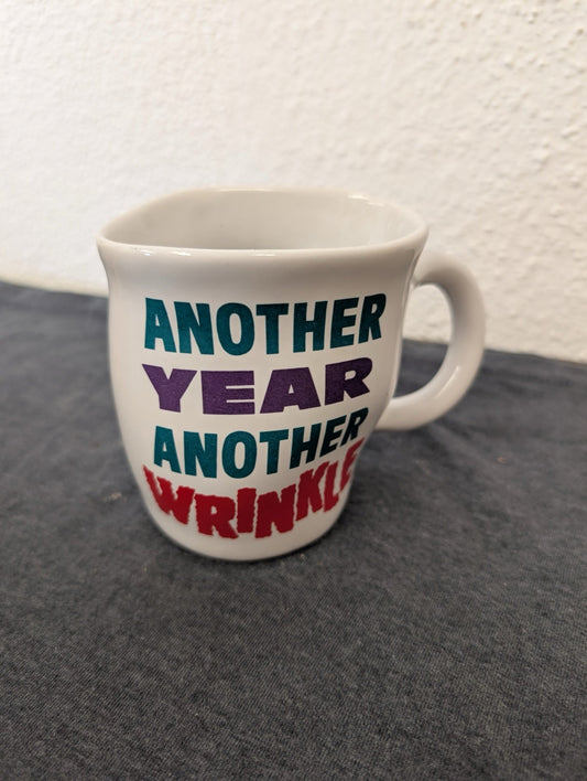 White coffee cup with the text Another Year Another Wrinkle in teal, purple, and red.
