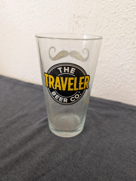 Clear pint beer glass featuring The Traveler Beer Co logo and a stylized drawn mustache.
