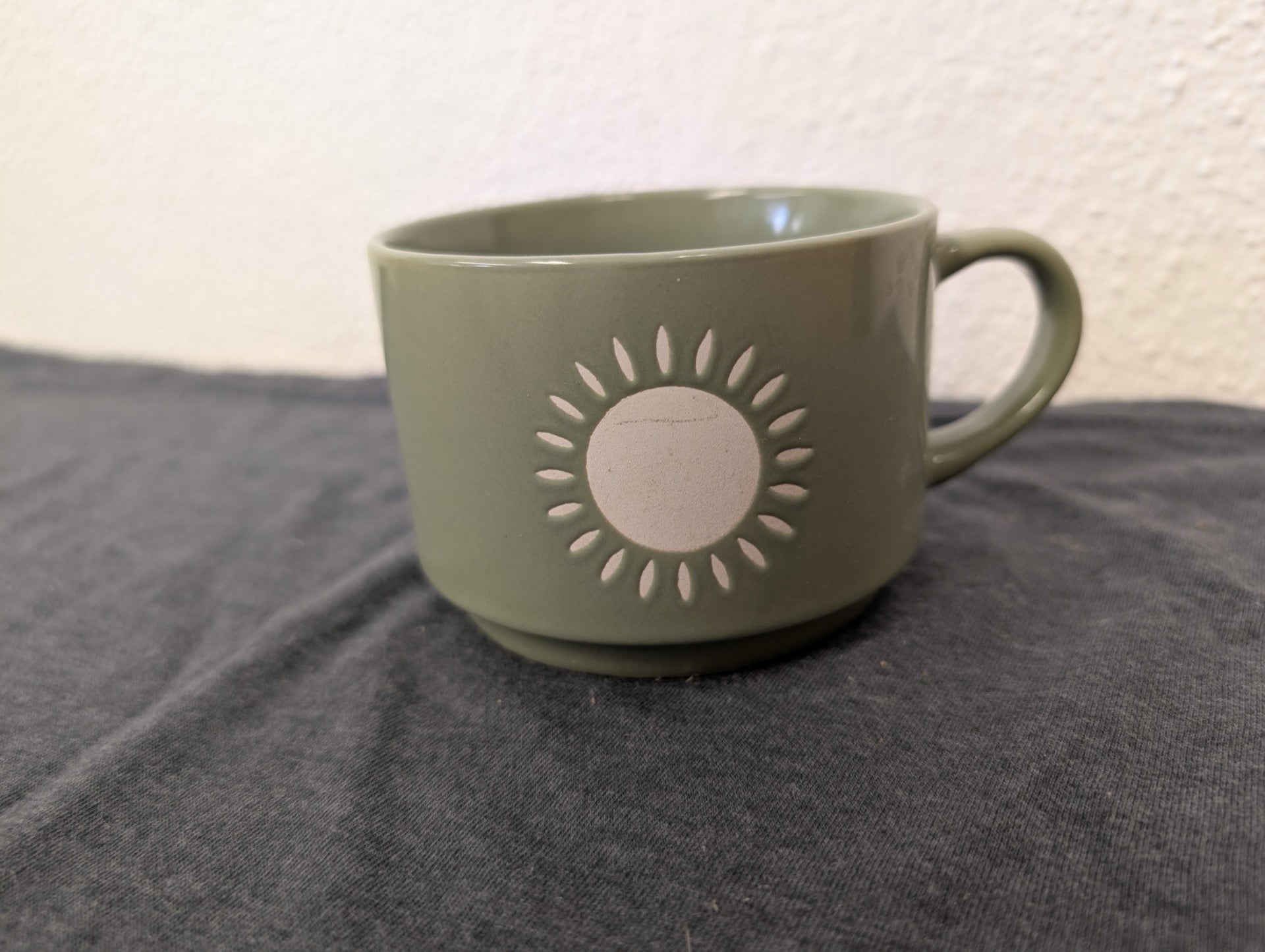 Light olive green ceramic coffee mug with a white circular sunburst design.