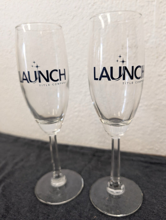 Two clear champagne flutes with the Launch Title Company logo printed in dark blue.