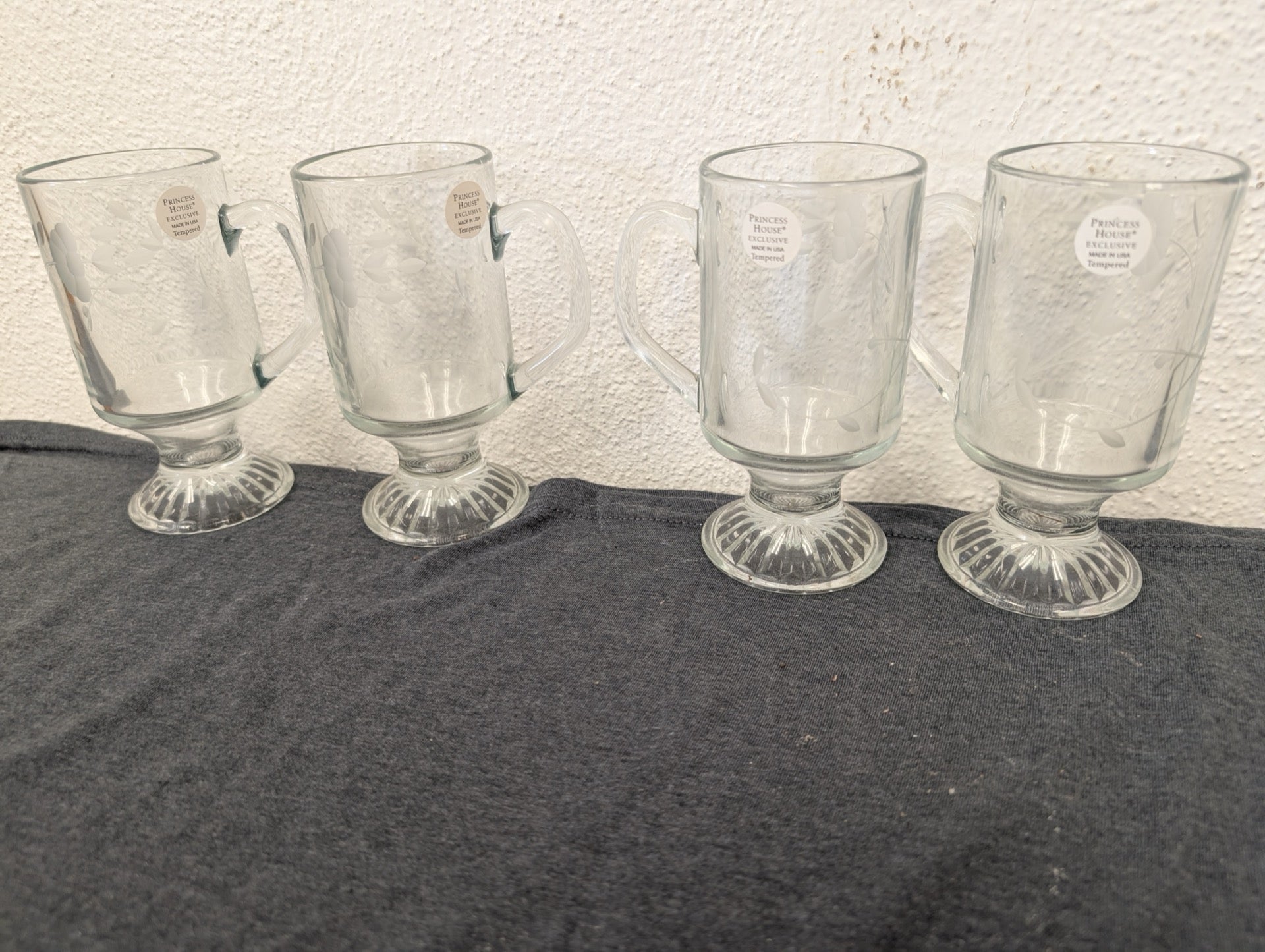 Four clear glass pedestal mugs lined up, featuring delicate floral etching and foil stickers.