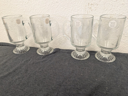 Four clear glass pedestal mugs lined up, featuring delicate floral etching and foil stickers.