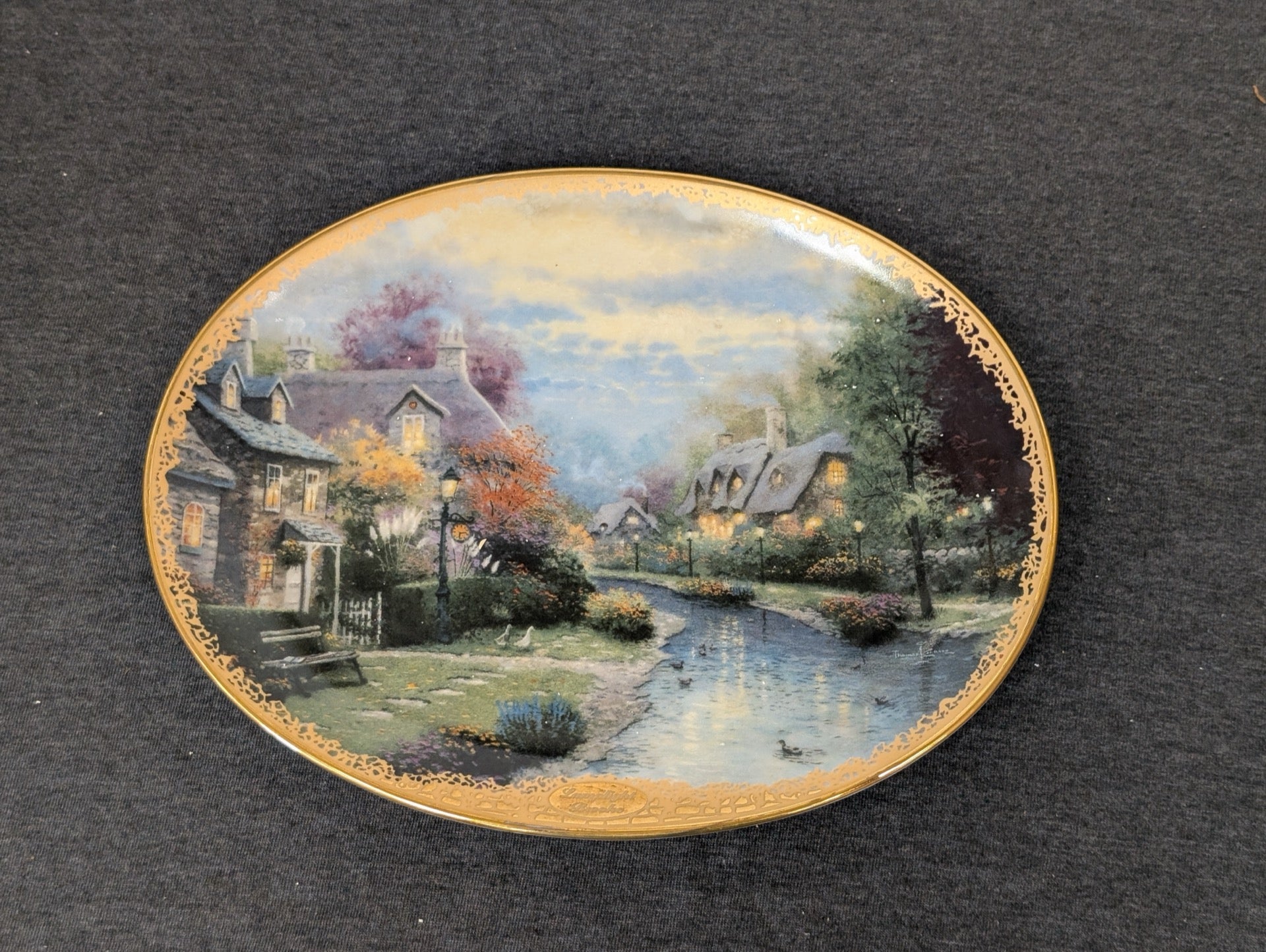 Oval porcelain plate depicting a glowing cottage beside a stream by Thomas Kinkade.