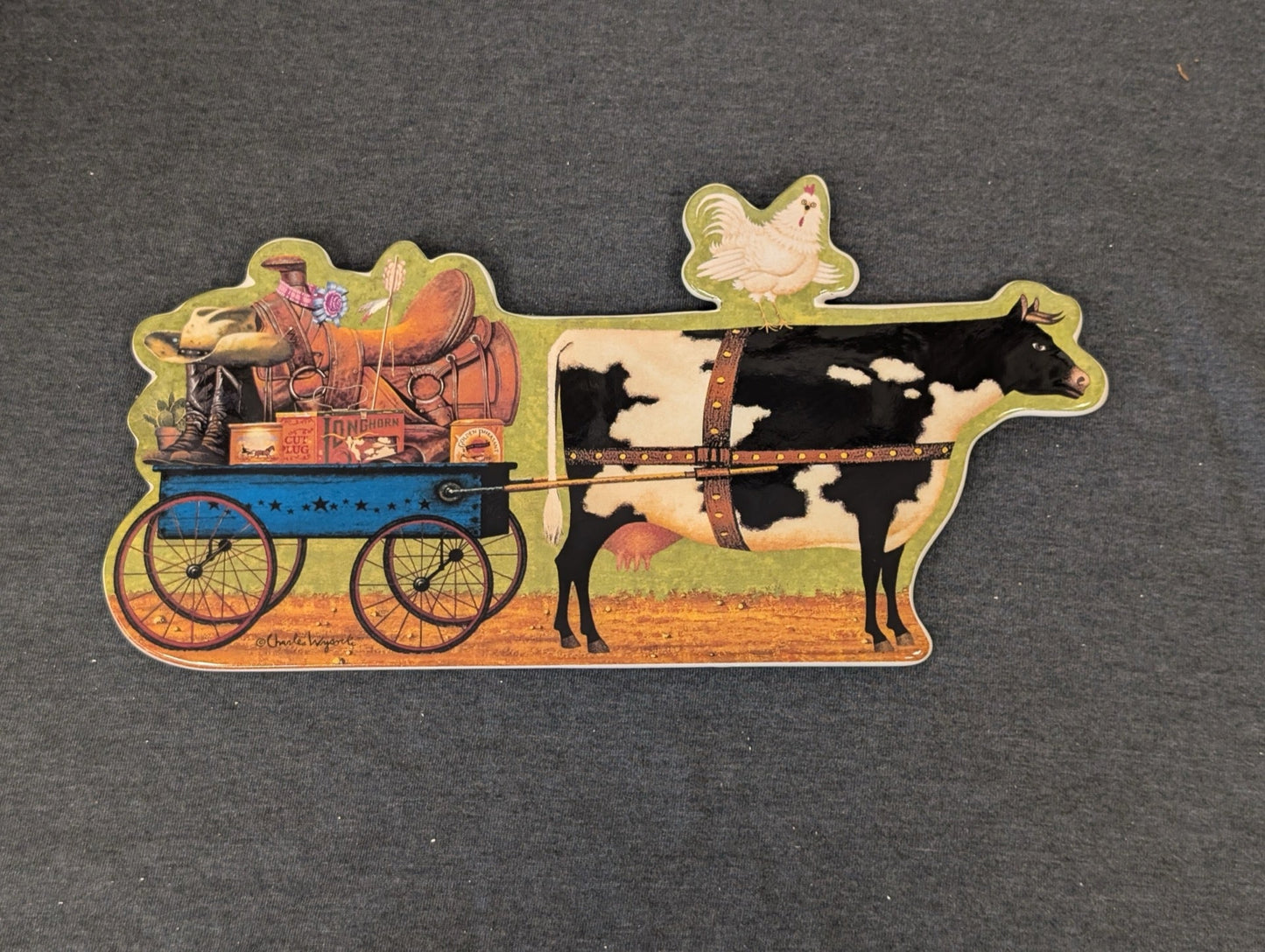 Shaped ceramic plaque of a black and white cow pulling a wagon, with a chicken on its back.