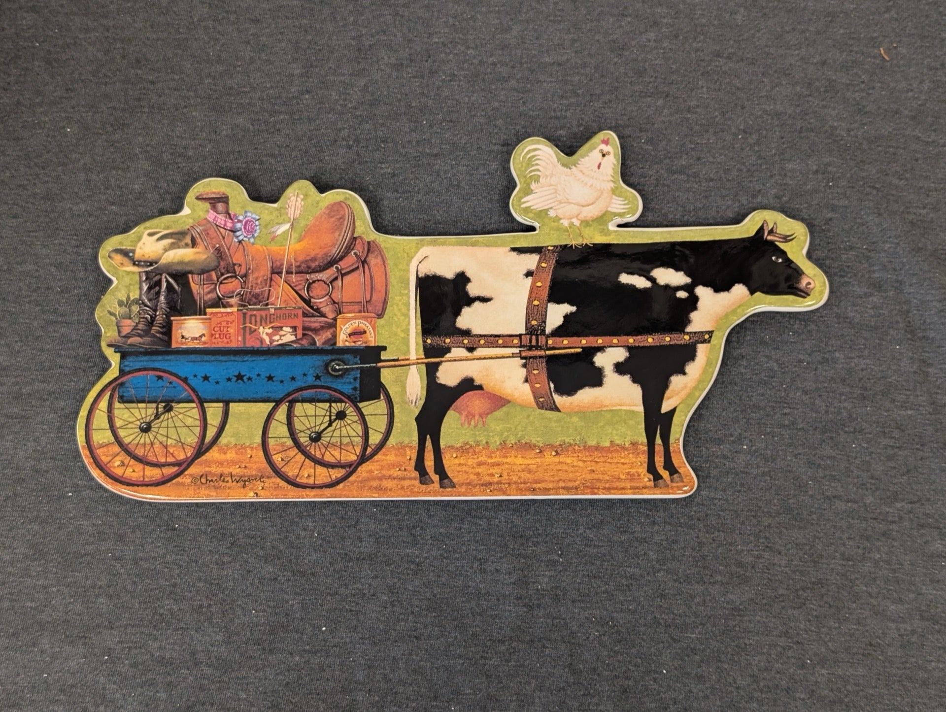 Shaped ceramic plaque of a black and white cow pulling a wagon, with a chicken on its back.