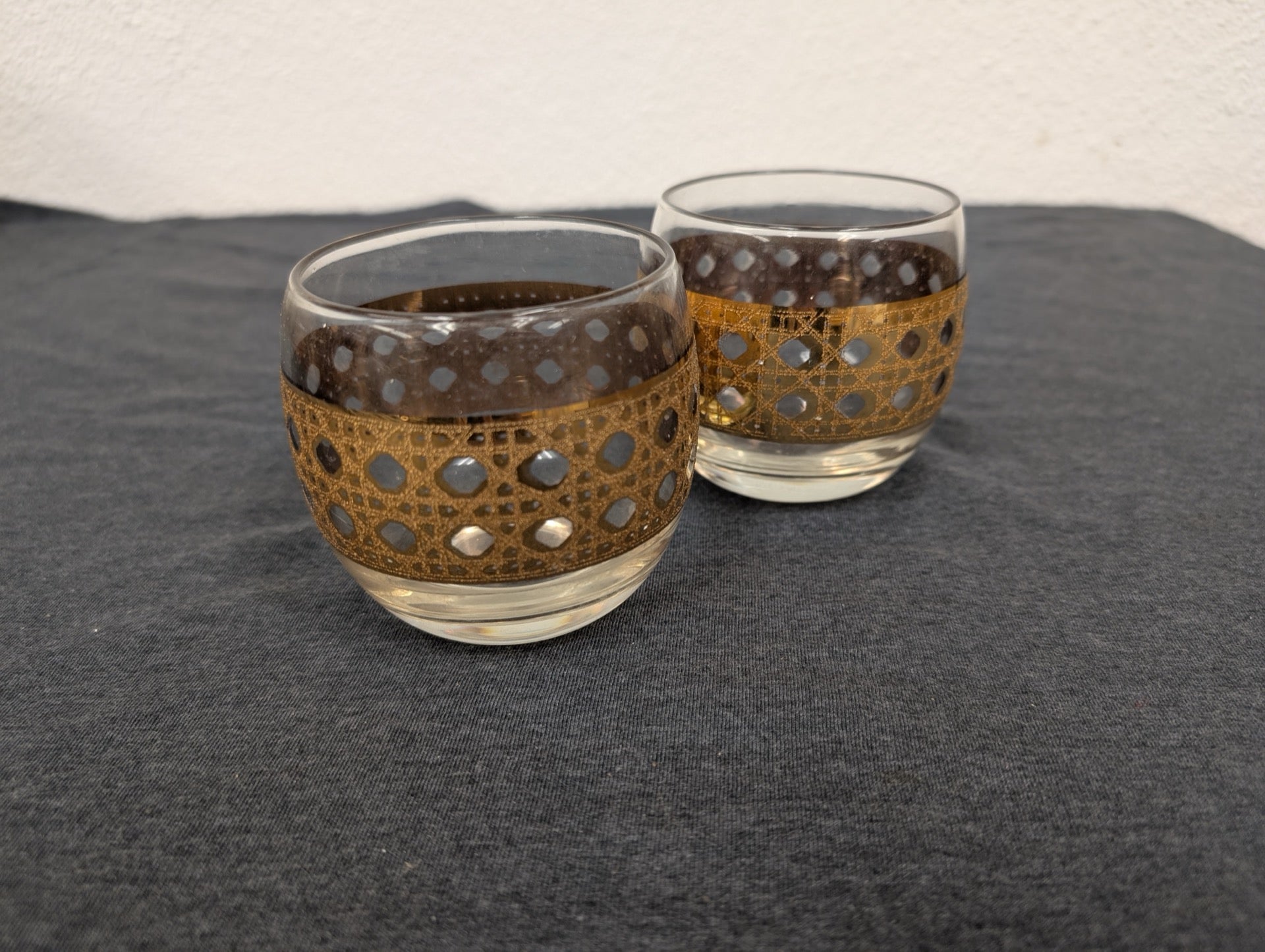 Two lowball clear glass cups with a thick geometric gold filigree band.