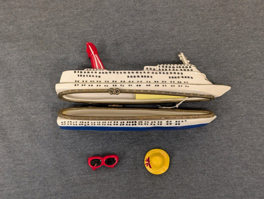 Open ceramic trinket box shaped like a cruise ship, with a tiny red sunglasses and yellow hat beside it.