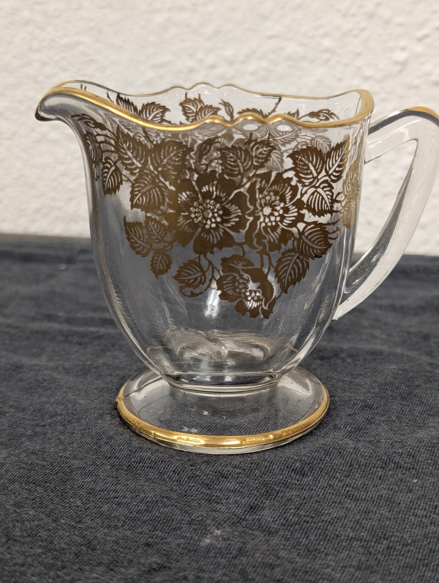 Small clear glass creamer pitcher with an intricate gold floral pattern and gold rim.