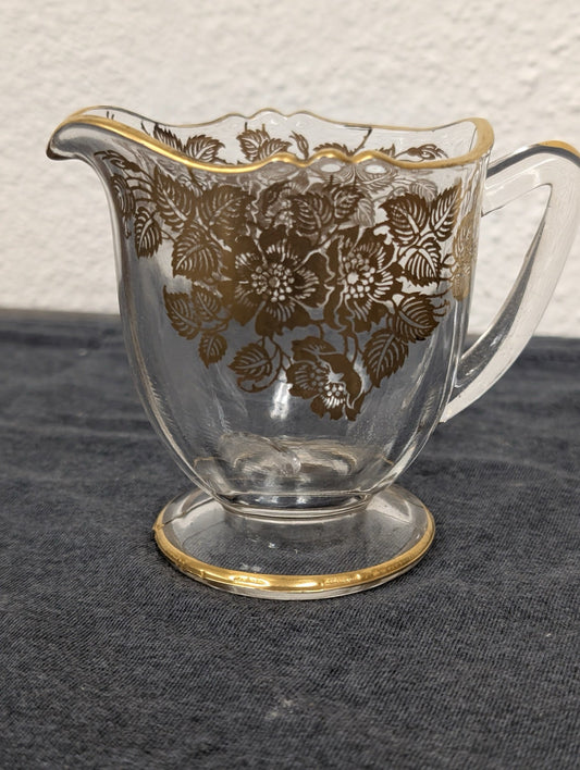 Small clear glass creamer pitcher with an intricate gold floral pattern and gold rim.
