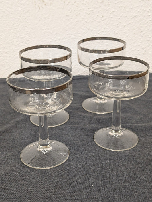 Four clear glass coupe glasses with thick silver rims arranged on a dark surface.