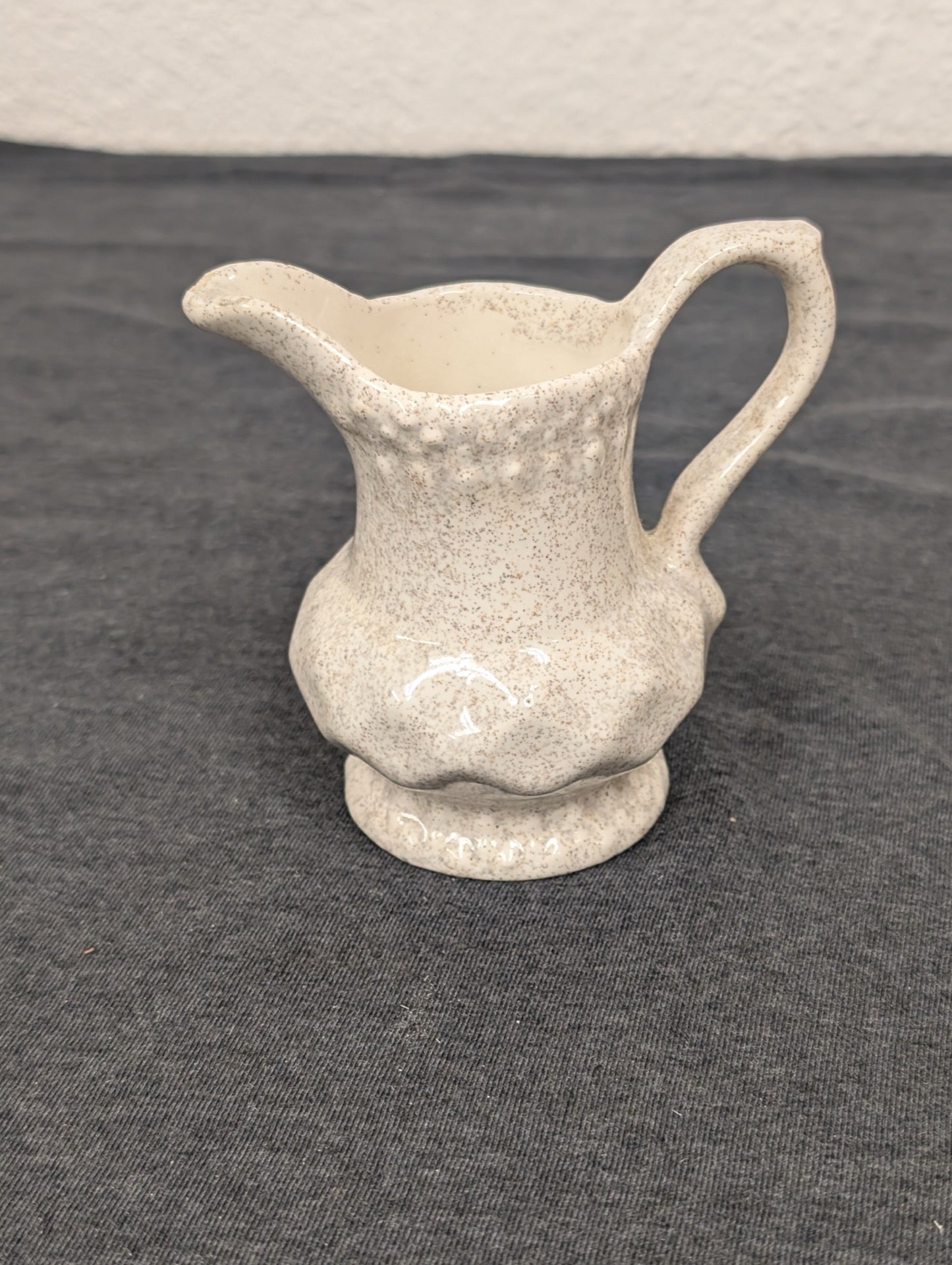 Small ceramic creamer pitcher with a beige speckled glaze and a scalloped bottom edge.