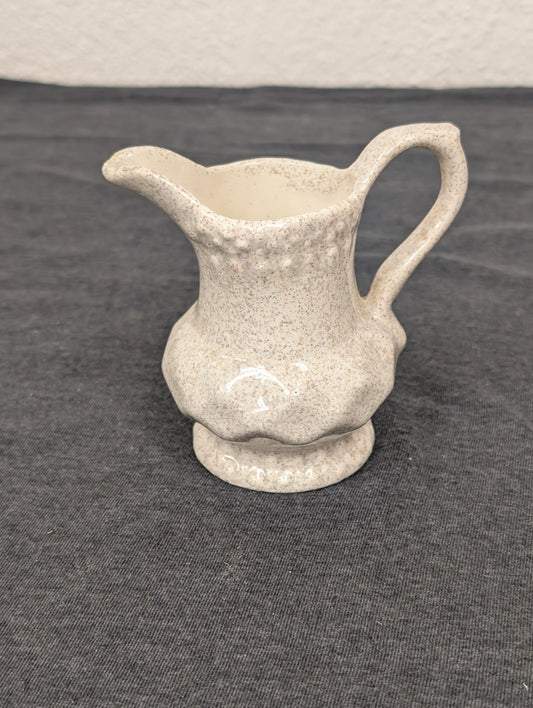 Small ceramic creamer pitcher with a beige speckled glaze and a scalloped bottom edge.