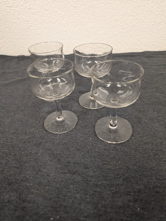Four small clear glass stemmed cordial glasses arranged on a dark surface.