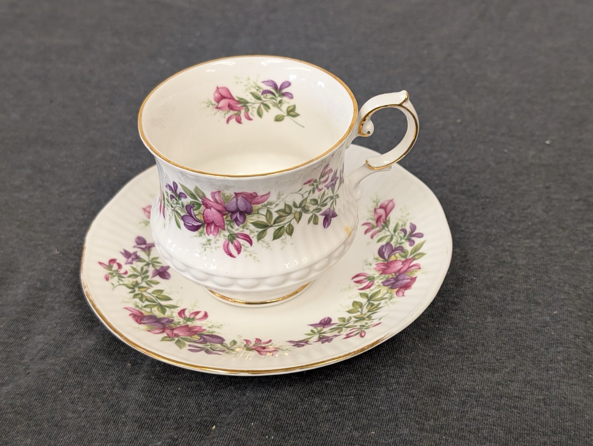 Teacup and saucer set featuring purple and pink delicate flowers and green leaves with gold rim over white bone china.
