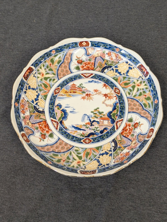Round decorative plate featuring intricate Asian Imari style designs with red and orange flowers, blue geometric patterns, and a central landscape.