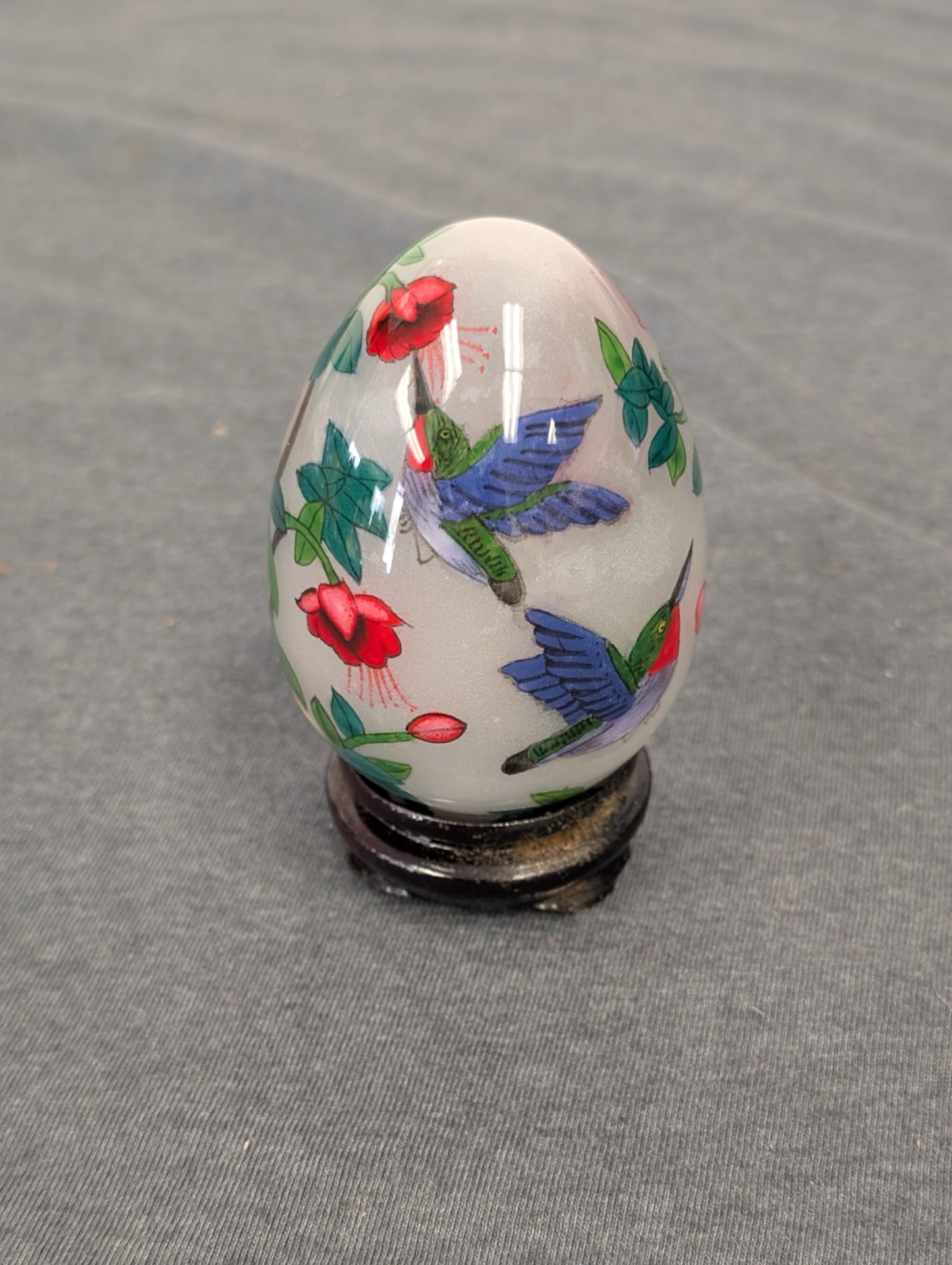 Frosted glass egg paperweight with painted hummingbirds and red flowers on a wooden stand.