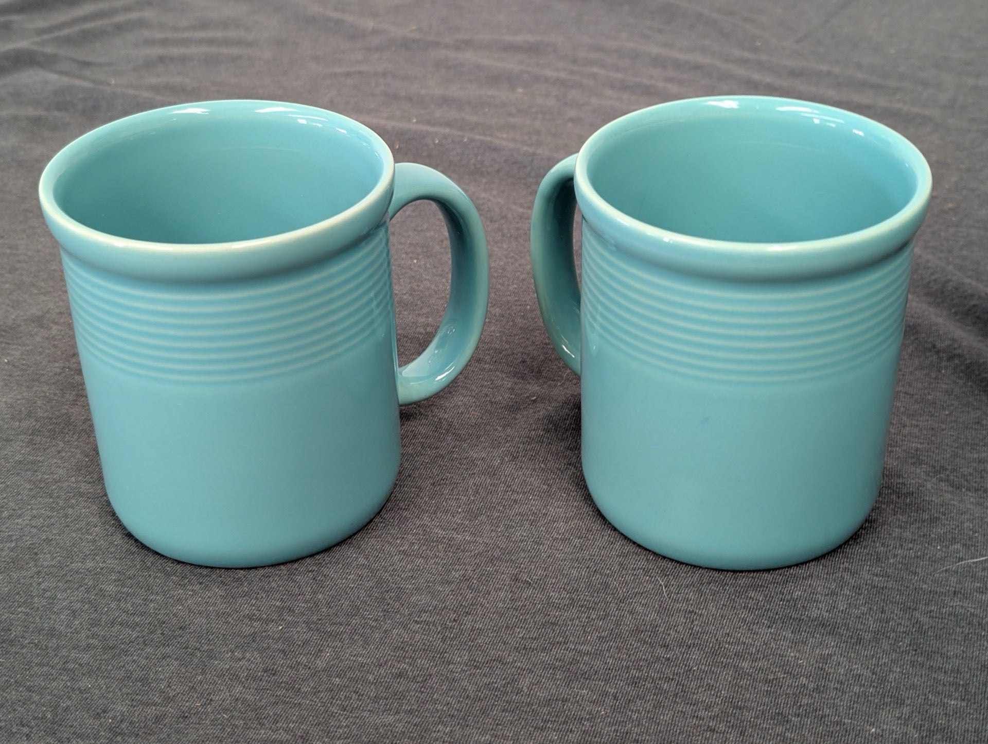 Two identical teal blue ceramic mugs with horizontal ribbed detailing on a gray background.