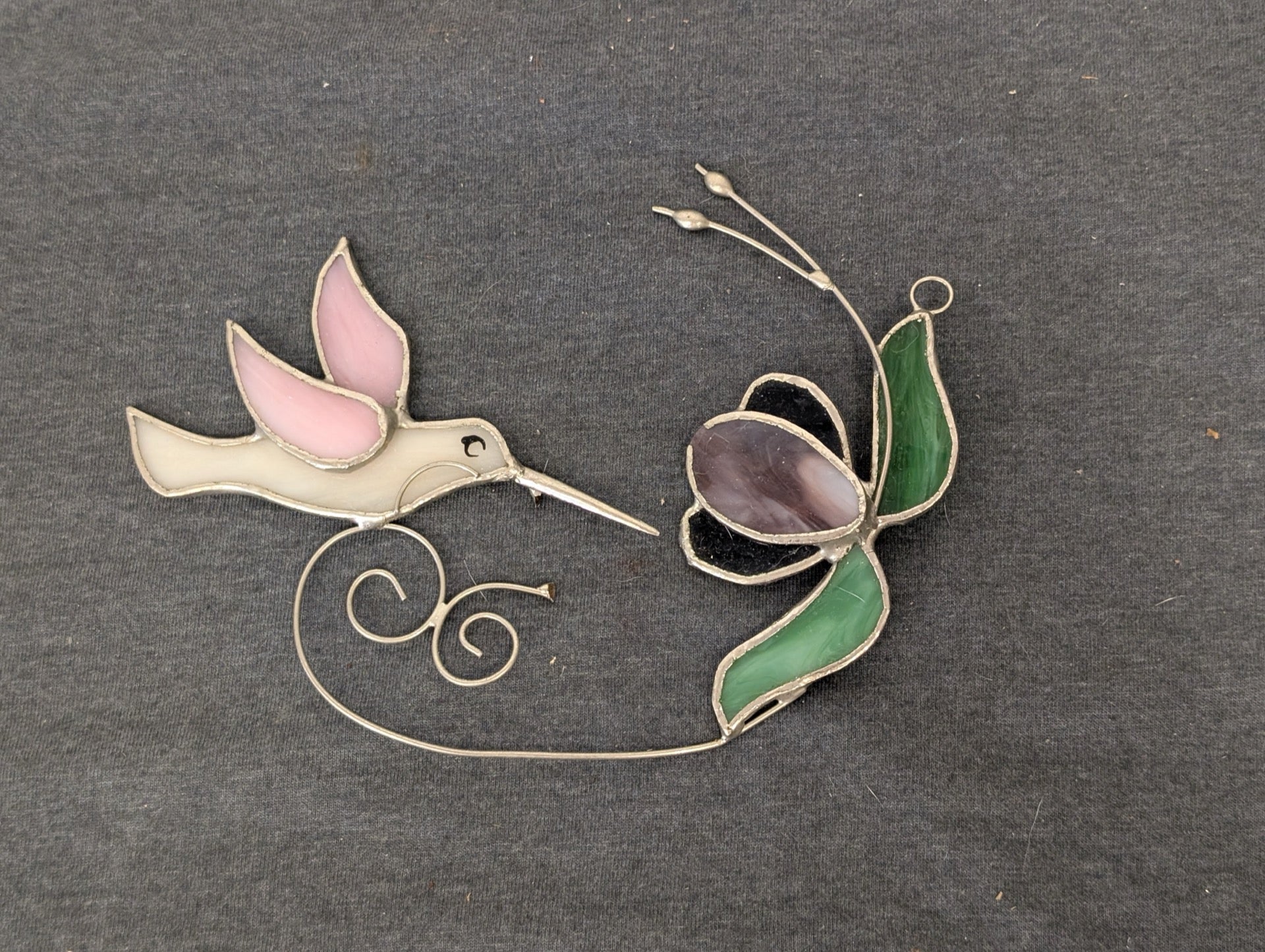 Stained glass art piece depicting a pink and white hummingbird and a purple flower connected by wire.