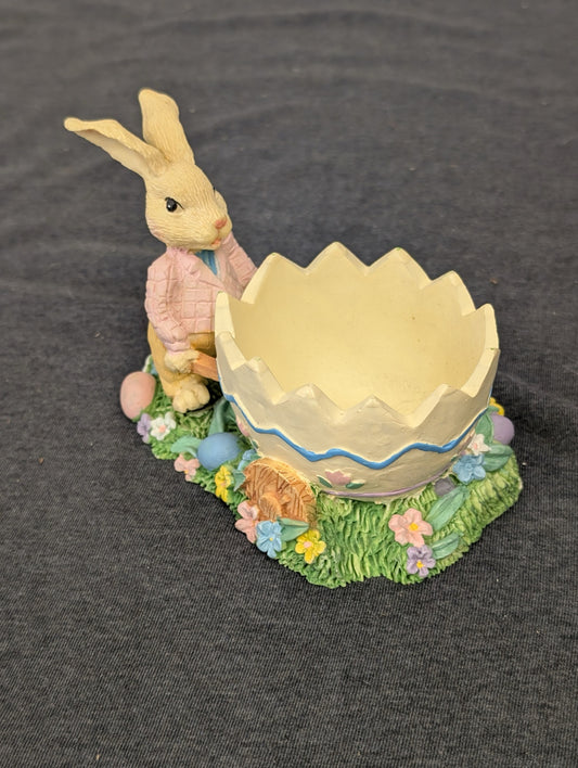 Easter bunny figurine in a pink jacket pushing a large broken eggshell wheelbarrow.